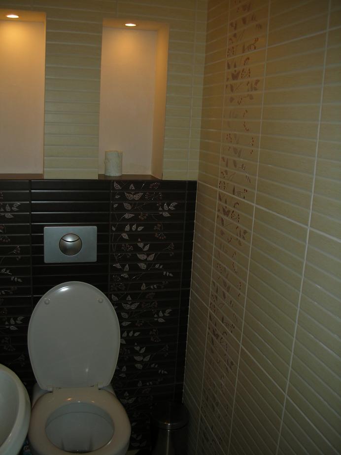 Ukończona łazienka z jasnymi, poziomymi płytkami na ścianie i ciemnymi płytkami z motywem roślinnym za toaletą; widoczne wnęki z oświetleniem.