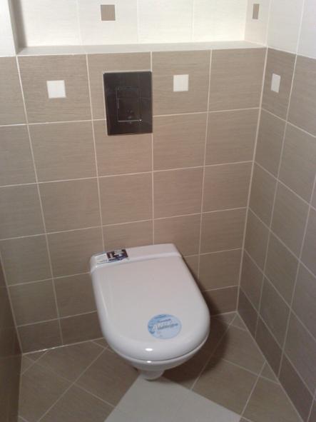 Beżowe płytki kwadratowe z białymi wstawkami w wykończonej łazience, wisząca toaleta z czarnym przyciskiem spłukiwania, widok z dołu.