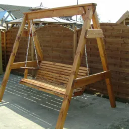 Drewniana huśtawka ogrodowa z łańcuchami, widoczna na tle drewnianego płotu i elementów architektury ogrodowej.