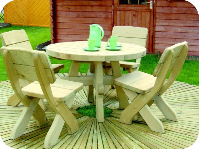 Drewniany okrągły stół z czterema krzesłami na tarasie z desek, zielony dzbanek i filiżanki na stole, drewniany domek ogrodowy w tle.