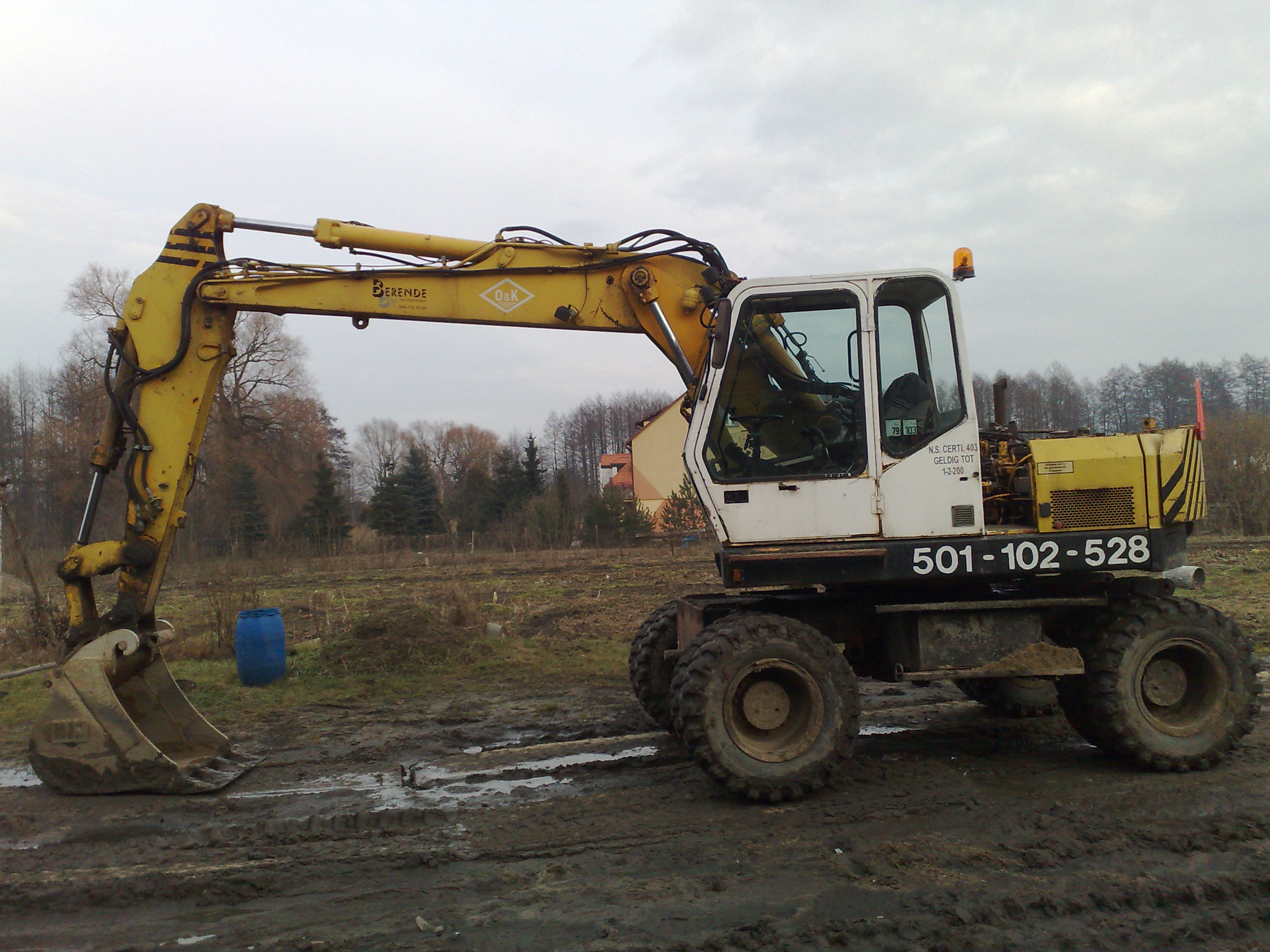 Żółto-biała koparka kołowa na błotnistym terenie w Warszawie, widoczny operator w kabinie i niebieski pojemnik w tle.