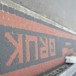 Ułożona kostka brukowa w kolorze czerwonym i szarym tworząca napis 'BRIK' wzdłuż krawędzi chodnika. Widok z góry, z fragmentem asfaltu w tle.