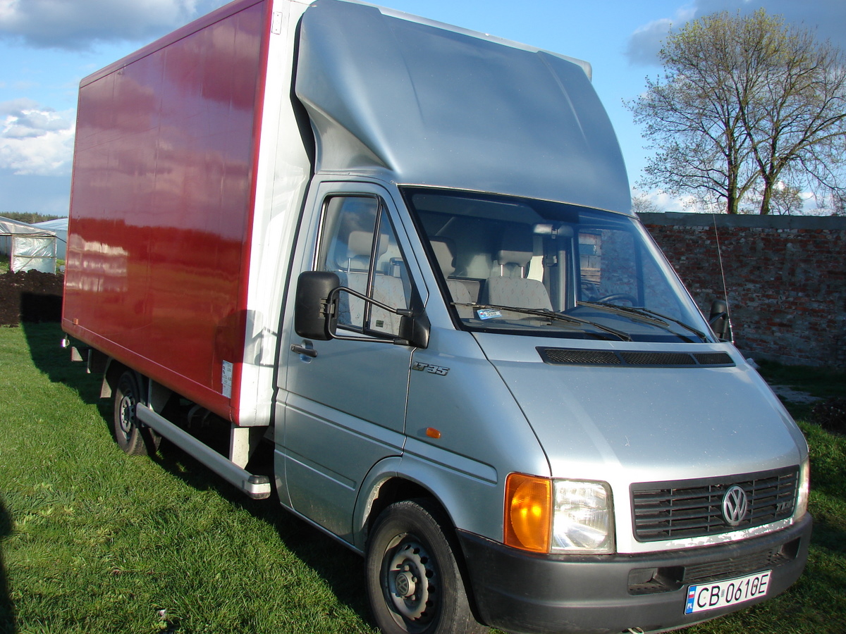 Srebrny samochód dostawczy Volkswagen LT35 z czerwoną zabudową kontenerową stoi na trawie przed ceglanym murem i szklarnią w tle.