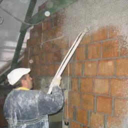 Pracownik w ubraniu roboczym nanosi zaprawę tynkarską na ścianę z cegły za pomocą maszyny natryskowej, widoczne fragmenty instalacji elektrycznej.