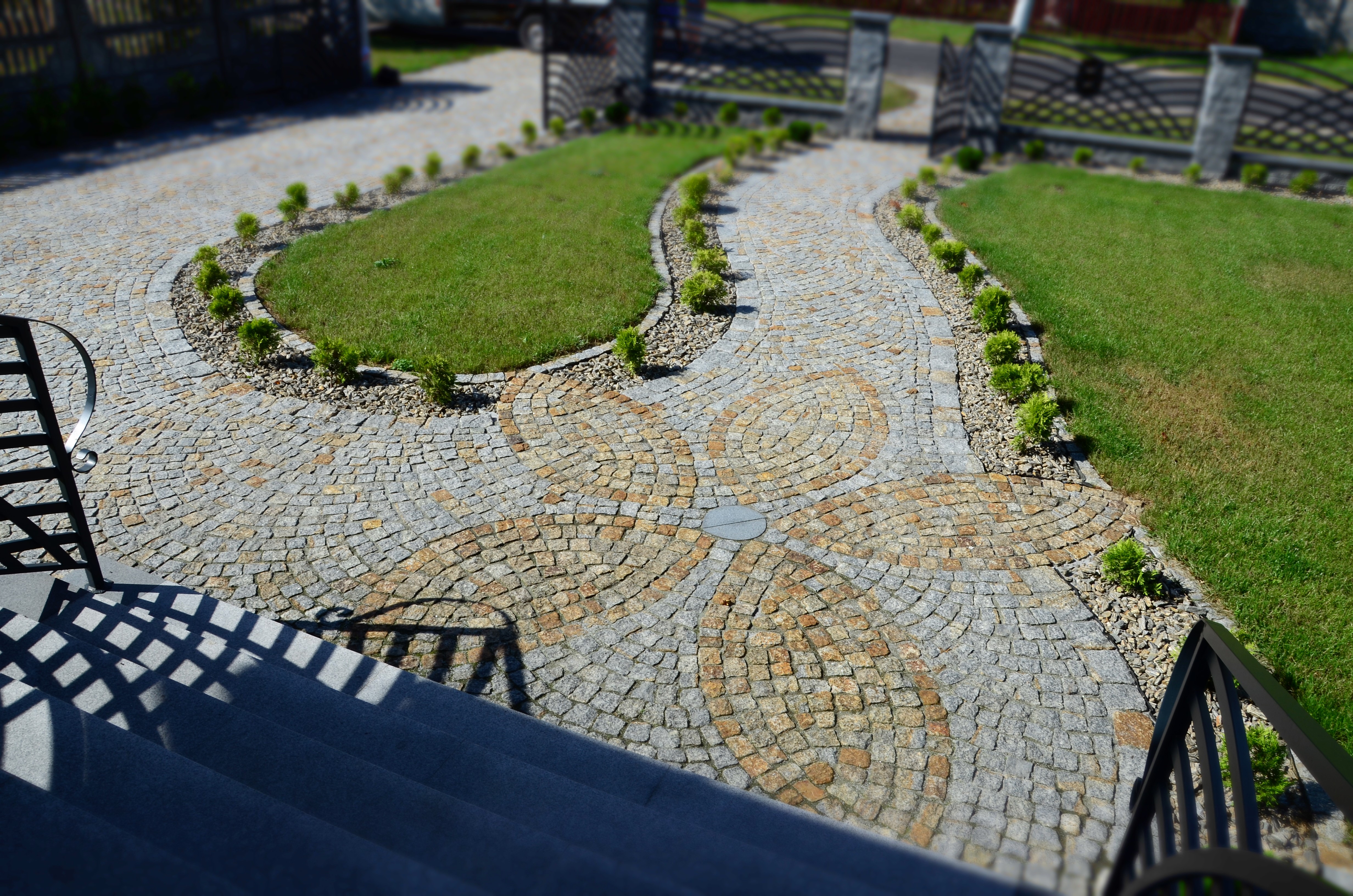 Ogród z krętą ścieżką z kostki brukowej ułożonej we wzór kwiatu, obrzeża z kamyków i niskich krzewów, widok z góry schodów.