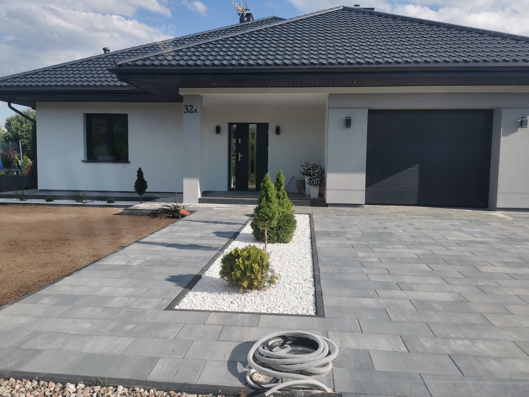 Szara kostka brukowa prowadząca do nowoczesnego domu z białymi ścianami i ciemną dachówką, ozdobiona białym żwirem i niskimi krzewami, zwinięty szary wąż leży na kostce.