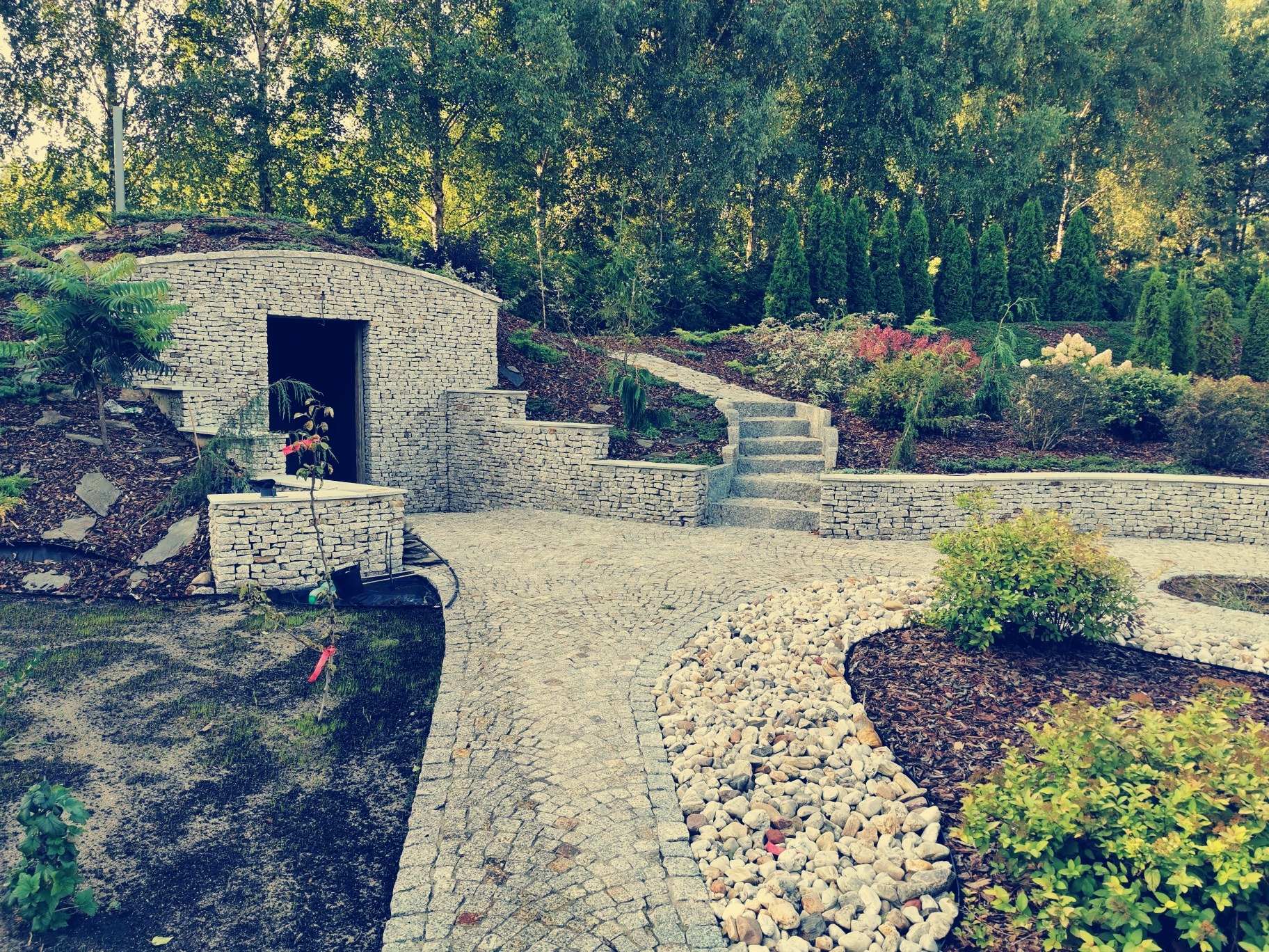 Ogród z kamienną ścieżką, schodami i piwniczką ziemną z kamienia, otoczony zielenią i drzewami.