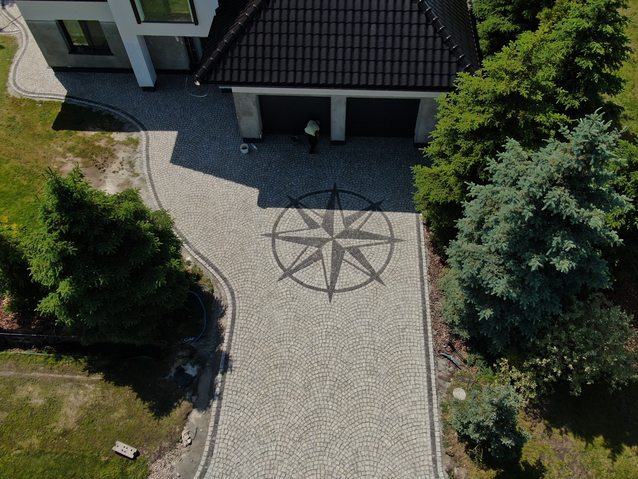 Widok z góry na podjazd z kostki brukowej ułożonej w wachlarz, z dekoracyjnym motywem róży wiatrów pośrodku, prowadzący do garażu i domu otoczonych zielenią