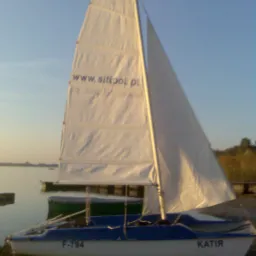 Biało-niebieska łódka żaglowa typu katamaran o nazwie 'Katia' z numerem F-794, zacumowana na brzegu jeziora, z widocznym żaglem z napisem 'www.sitipol.pl'.