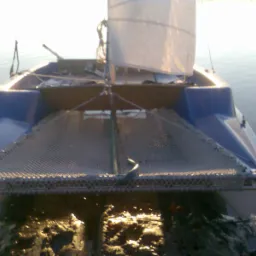 Niebieski katamaran z żaglem, widziany od przodu, częściowo zanurzony w wodzie i błocie, z widoczną siatką między pływakami.