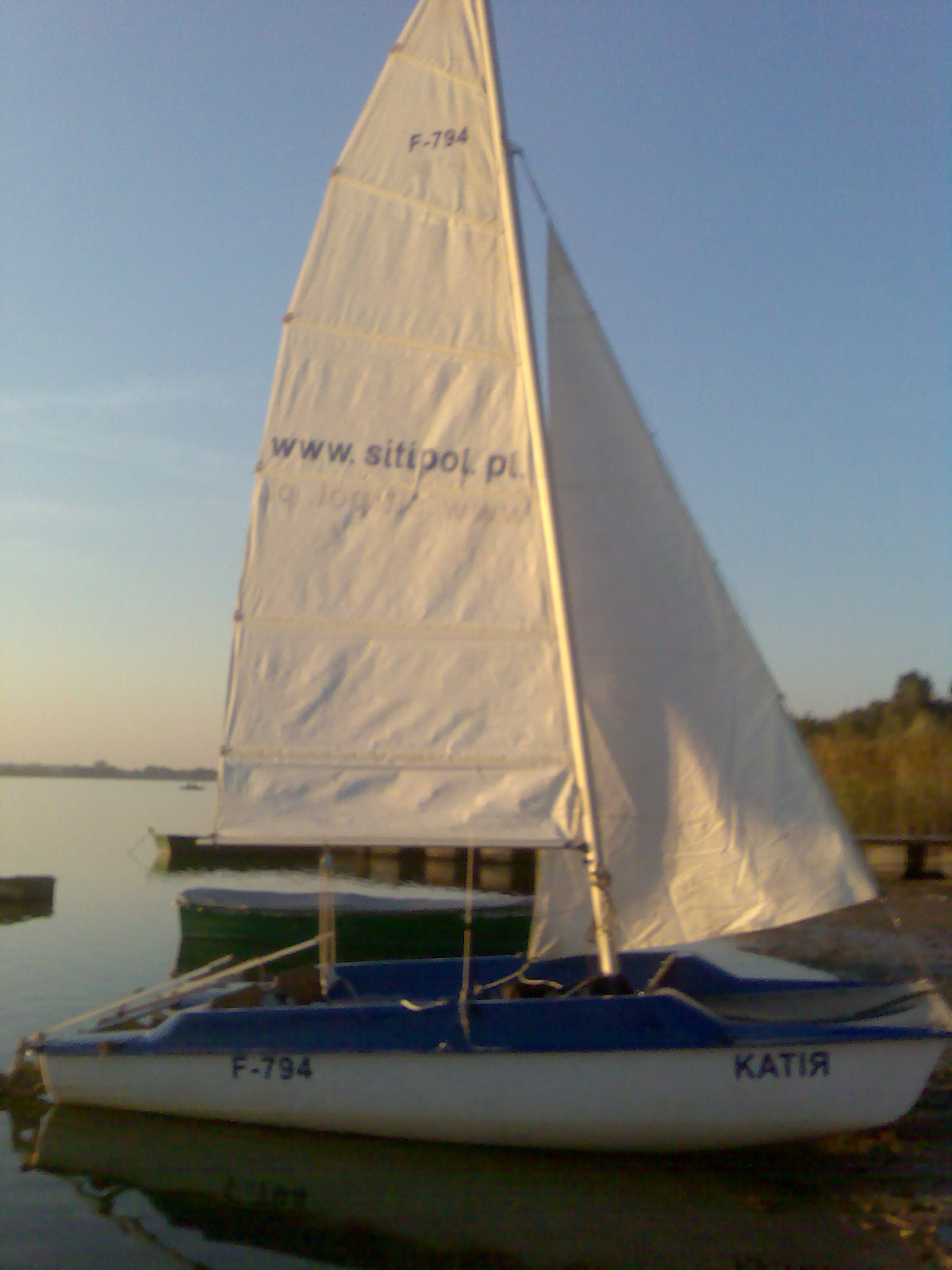 Biało-niebieska łódka żaglowa typu katamaran o nazwie 'Katia' z numerem F-794, zacumowana na brzegu jeziora, z widocznym żaglem z napisem 'www.sitipol.pl'.
