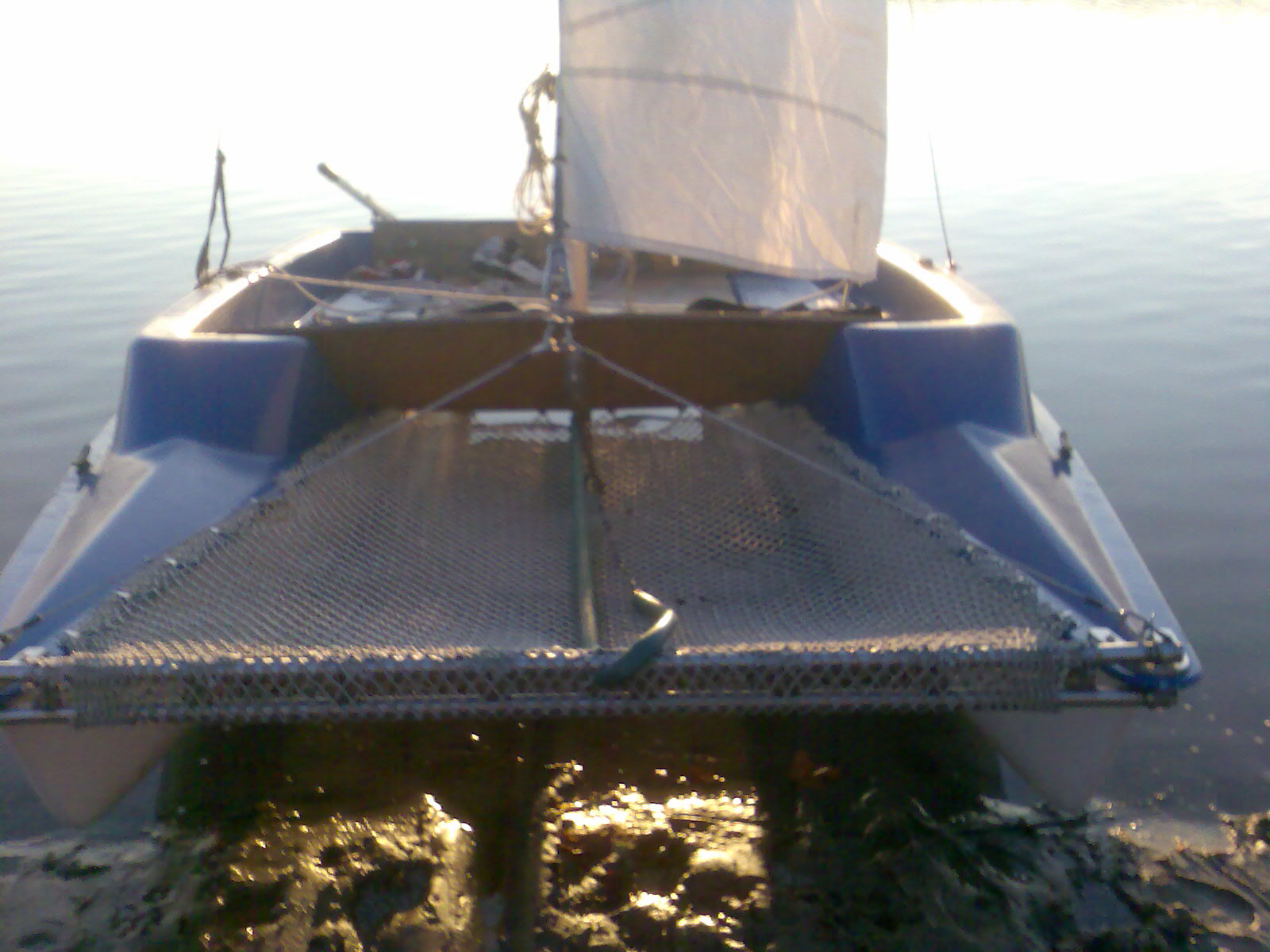 Niebieski katamaran z żaglem, widziany od przodu, częściowo zanurzony w wodzie i błocie, z widoczną siatką między pływakami.