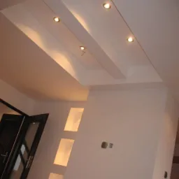 Biały sufit podwieszany z wbudowanymi lampami punktowymi, oświetlona wnęka w ścianie i częściowo widoczne drzwi z szybami.