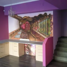 Fioletowe wnętrze z biurkiem z jasnego drewna, nad którym znajduje się abstrakcyjny mural na ścianie, oraz kryształowy żyrandol.