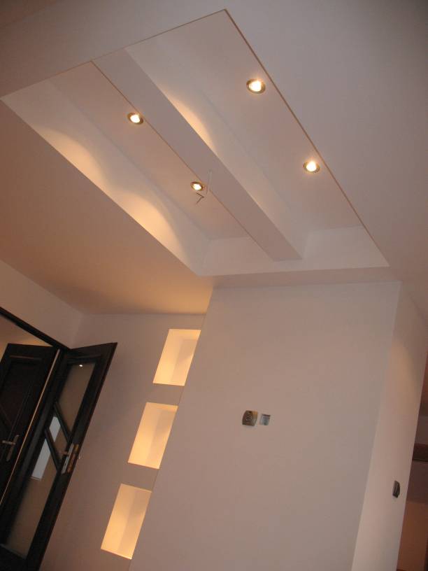 Biały sufit podwieszany z wbudowanymi lampami punktowymi, oświetlona wnęka w ścianie i częściowo widoczne drzwi z szybami.