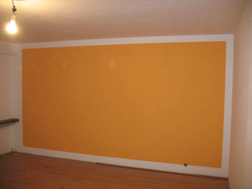 Świeżo pomalowana na pomarańczowo ściana z białym obramowaniem w pustym pokoju z parkietem i żarówką na suficie.