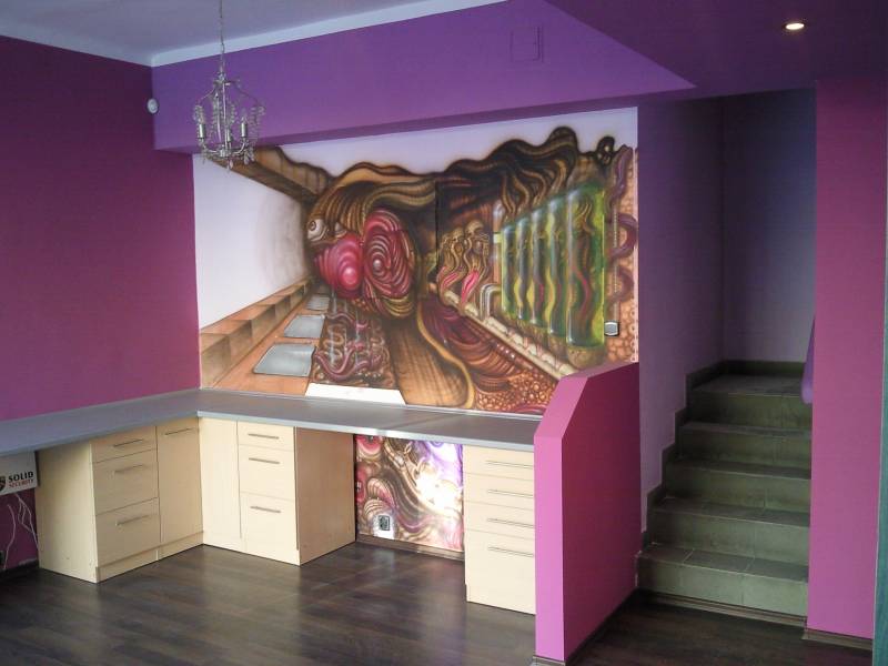 Fioletowe wnętrze z biurkiem z jasnego drewna, nad którym znajduje się abstrakcyjny mural na ścianie, oraz kryształowy żyrandol.