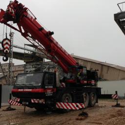 Uslugi dzwigowe Kielce - Czerwony dźwig samochodowy Grove na placu budowy, z wysięgnikiem skierowanym ku górze, w tle budynek przemysłowy i konstrukcja stalowa.