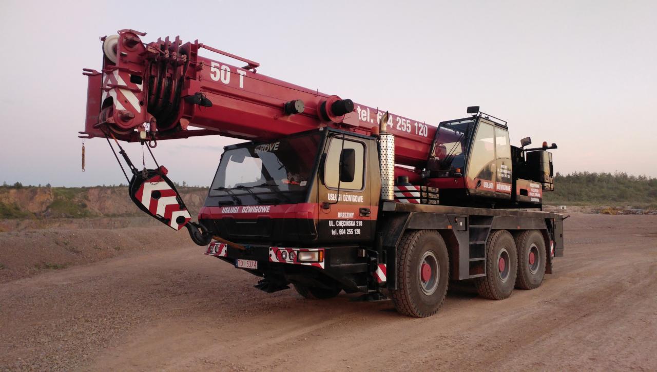 Czerwono-czarny dźwig samojezdny Grove o udźwigu 50 ton, z wysuniętym ramieniem, zaparkowany na żwirowym placu w pobliżu wyrobiska.