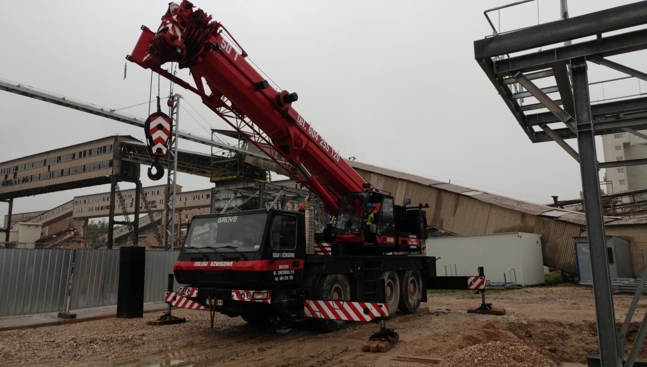 Czerwony dźwig samochodowy Grove na placu budowy, z wysięgnikiem skierowanym ku górze, w tle budynek przemysłowy i konstrukcja stalowa.