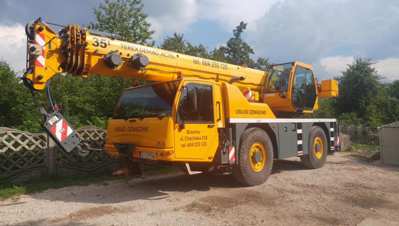 Żółty dźwig samochodowy Terex Demag AC35L z wysięgnikiem, zaparkowany na żwirowym terenie, z widocznym adresem firmy i numerem telefonu na kabinie.