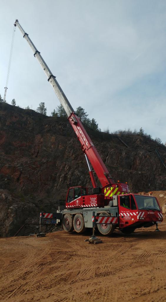 Czerwony, terenowy dźwig samochodowy Terex z wysuniętym ramieniem na tle skalistego wzgórza i pochmurnego nieba. Dźwig stoi na ziemi z wyraźnymi śladami opon.