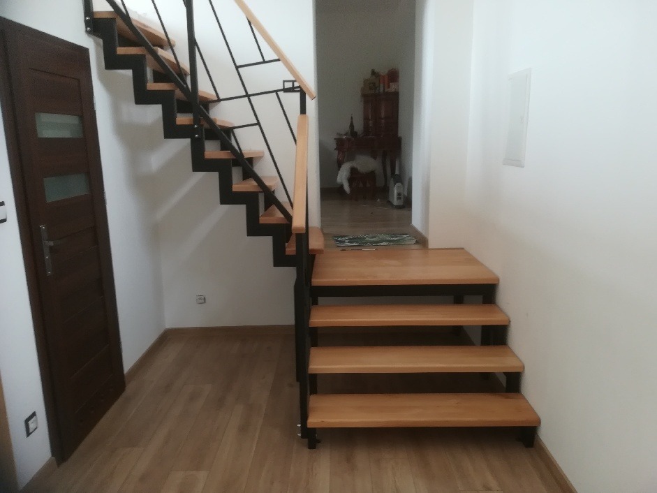 Drewniane schody z czarną metalową konstrukcją i drewnianą barierką, widok z dołu na korytarz.