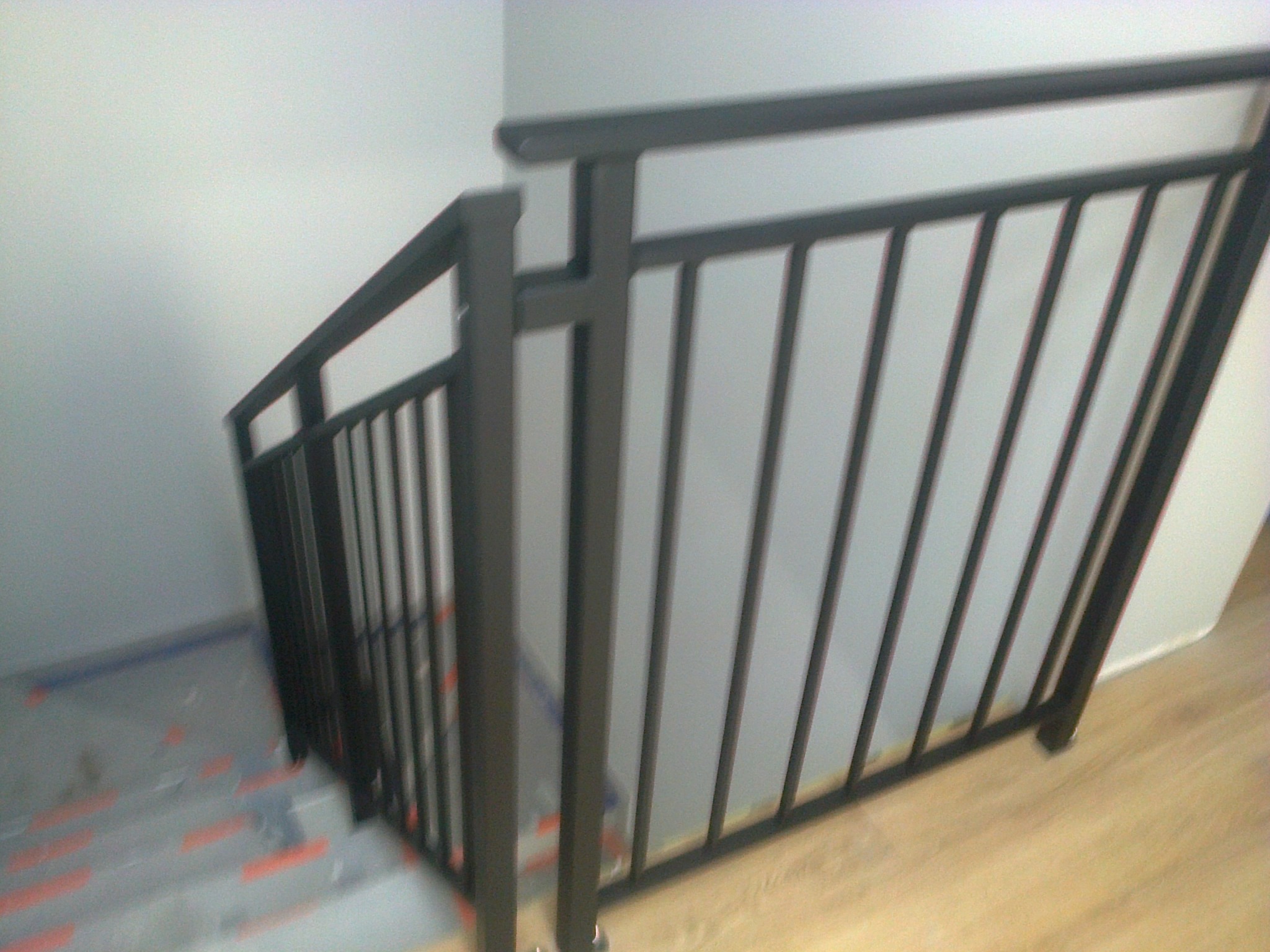 Nowoczesna, czarna balustrada schodowa z pionowymi prętami, zamontowana na schodach zabezpieczonych folią malarską, widok z góry.