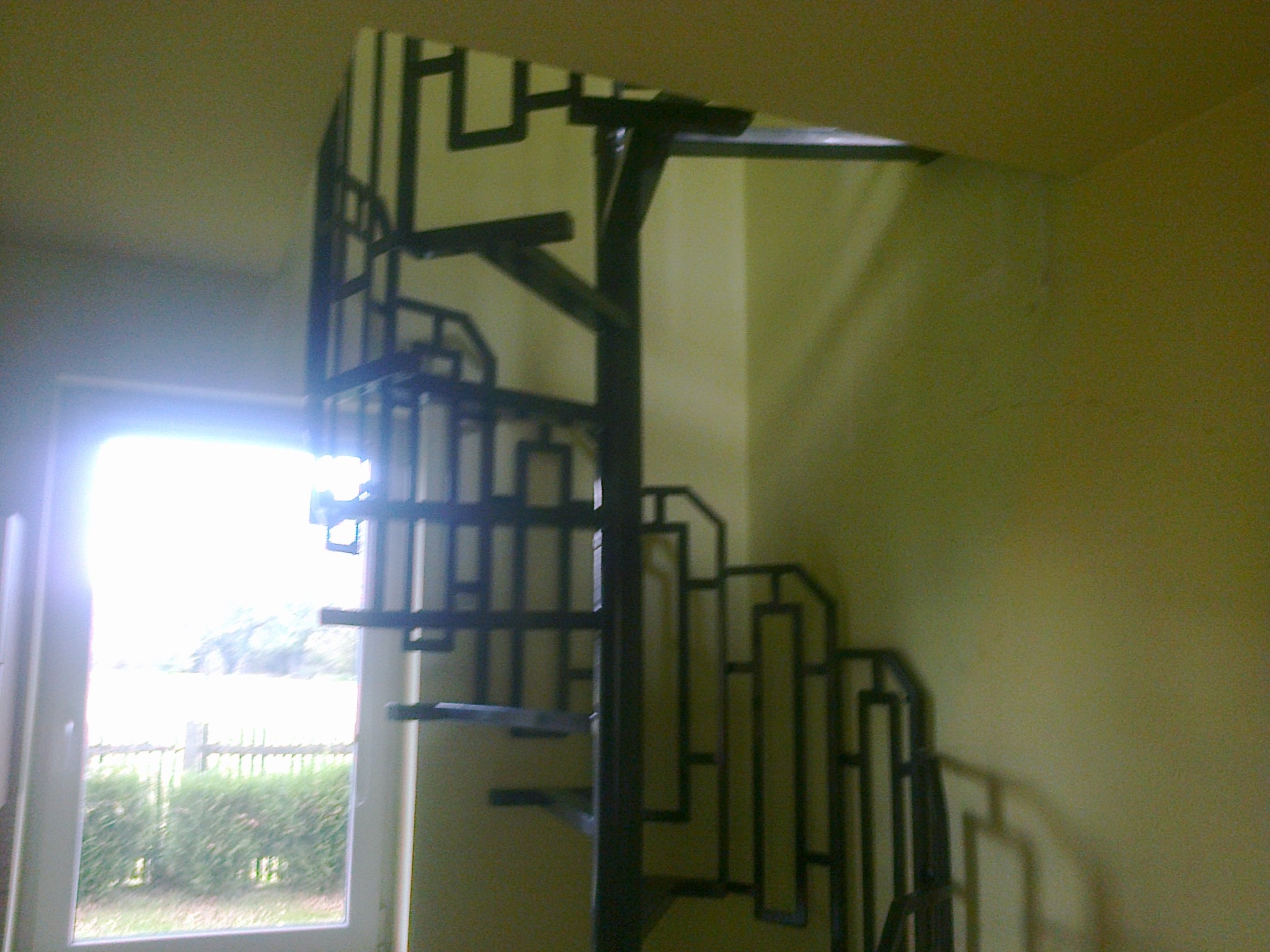 Metalowa konstrukcja ogrodzeniowa wewnątrz budynku, widoczna przez okno z fragmentem ogrodu i drewnianym ogrodzeniem.