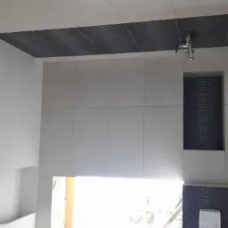 Nowoczesna łazienka z białymi płytkami, czarnym akcentem na suficie i wnęką z ciemną mozaiką, nietypowe wykończenie sufitu z falistym elementem dekoracyjnym.