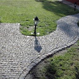 P.W.Le-trans - Ścieżka z drobnej kostki o nieregularnym kształcie, wijąca się przez trawnik, oświetlona latarnią. Widoczny fragment ceglanego muru budynku.