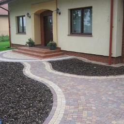 P.W.Le-trans - Kręta ścieżka z kostki brukowej w odcieniach brązu i beżu prowadząca do wejścia domu z niewielkimi schodami, obok rabaty wysypane korą.