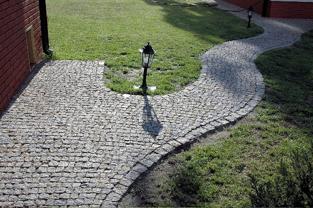 Ścieżka z drobnej kostki o nieregularnym kształcie, wijąca się przez trawnik, oświetlona latarnią. Widoczny fragment ceglanego muru budynku.