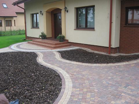 Kręta ścieżka z kostki brukowej w odcieniach brązu i beżu prowadząca do wejścia domu z niewielkimi schodami, obok rabaty wysypane korą.