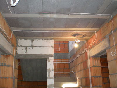 Surowy stan wnętrza budynku w trakcie budowy: ściany z czerwonej cegły i białych bloczków, sufit z betonowych płyt, widoczne przewody elektryczne prowadzone po ścianach i suficie, żarówka...