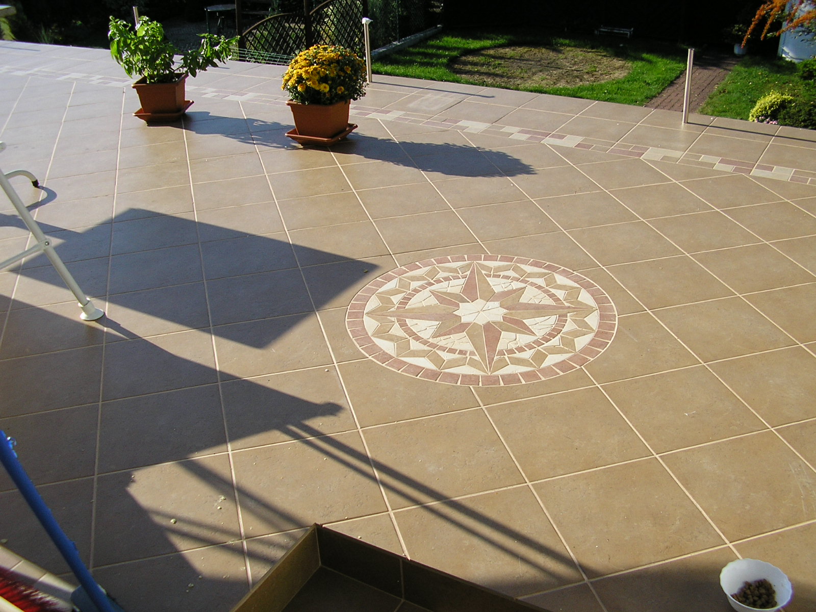 Beżowa, kwadratowa płytka ceramiczna na tarasie z dekoracyjnym motywem rozety w centrum, doniczki z roślinami w tle