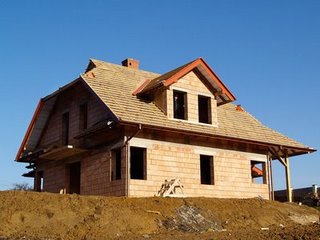 Surowy, dwukondygnacyjny dom w trakcie budowy z widoczną konstrukcją dachu pokrytą brązową dachówką ceramiczną, ściany z jasnych pustaków, brak okien, ziemia wokół budynku.