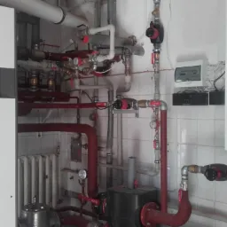 Kotłownia z dwoma kotłami ACV (HeatMaster i Prestige), licznymi rurami, zaworami, pompami obiegowymi, grzejnikiem oraz elementami instalacji elektrycznej na ścianie wyłożonej białymi płytkami.