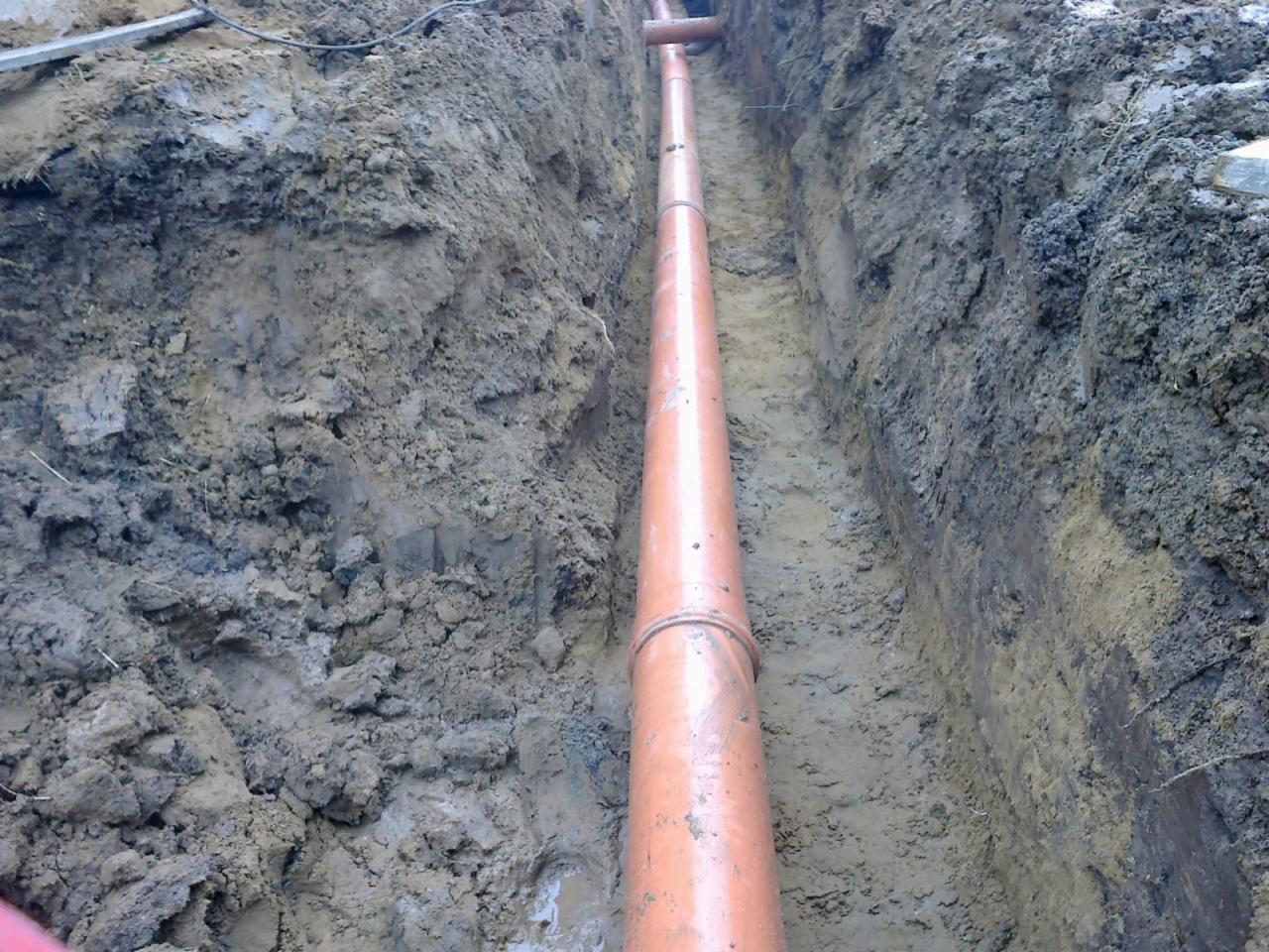 Pomarańczowa rura w wykopie ziemnym, widoczne ślady błota i wilgoci na ścianach wykopu, fragment drugiej rury w tle.