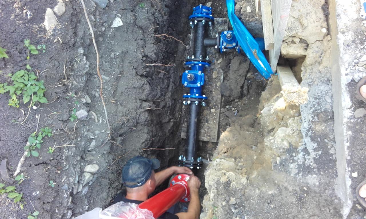 Instalacja rury wodociągowej w wykopie ziemnym, widoczne złącza kołnierzowe i zawór, pracownik łączy czerwoną rurę, zabezpieczenie wykopu deskami.