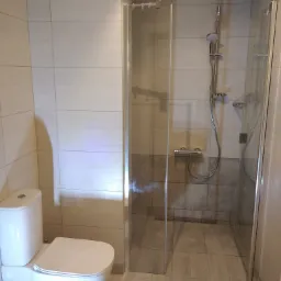 Wykończona łazienka z kabiną prysznicową z przezroczystego szkła, wiszącą słuchawką prysznicową i toaletą w kolorze białym, wyłożona płytkami w odcieniach beżu i szarości.