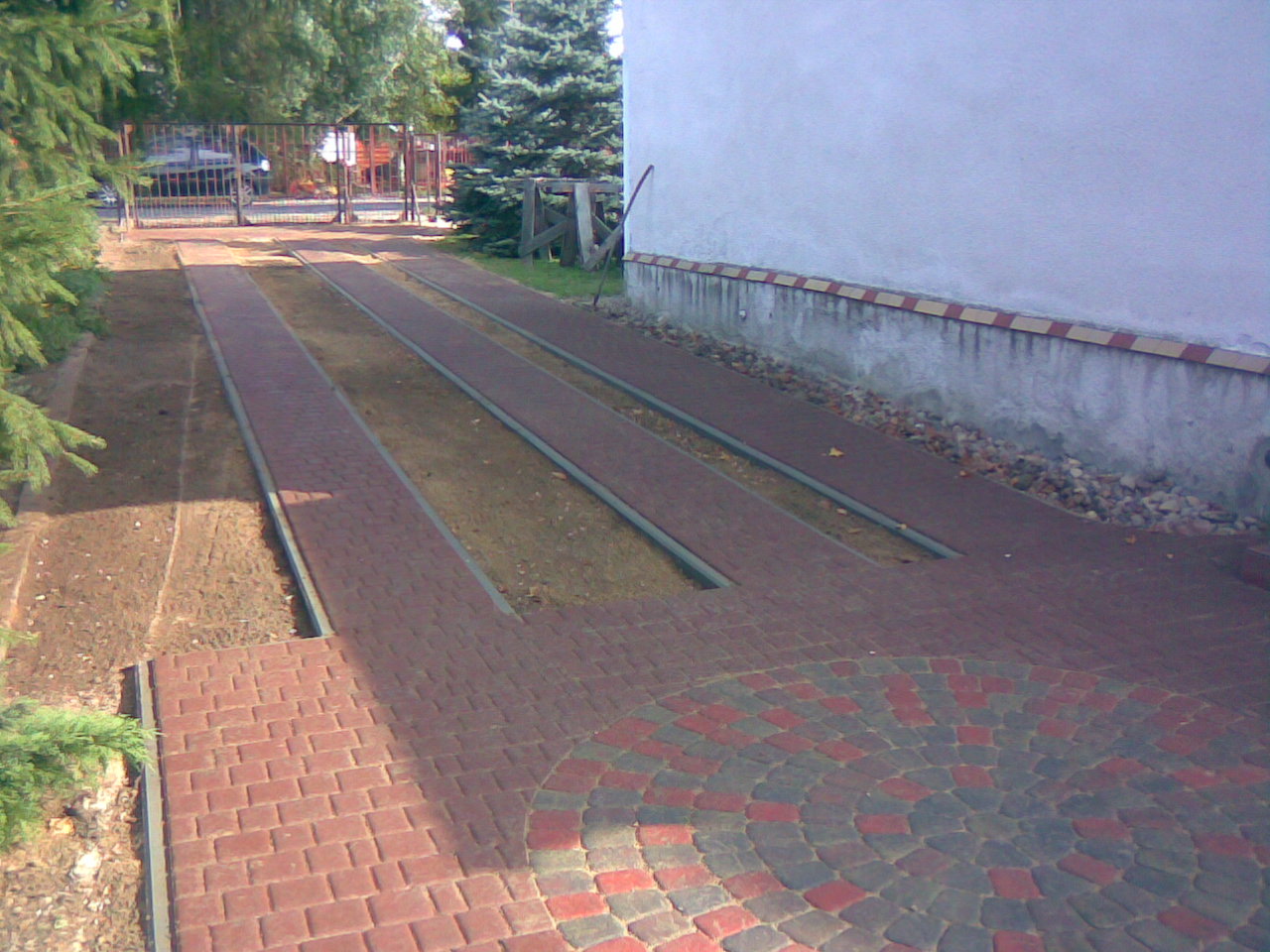 Częściowo ułożona czerwona kostka brukowa na podjeździe, widoczne przygotowane pasy ziemi, obrzeża i fragment okrągłego wzoru z czarno-czerwonej kostki.