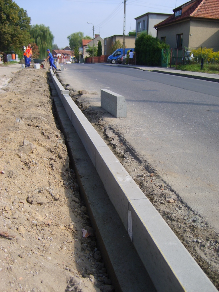 Montaż obrzeży chodnikowych z betonu na podsypce cementowej, widoczni pracownicy w odblaskowych ubraniach i niebieskim kombinezonie, ulica asfaltowa w tle.