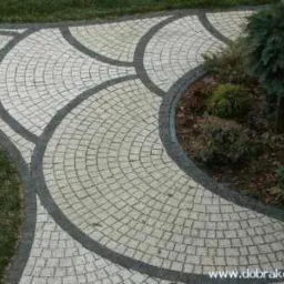 Ścieżka z jasnej kostki granitowej ułożona w wachlarze, obrzeża z ciemnej kostki, obok trawnik i rabata z krzewami.