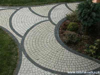 Ścieżka z jasnej kostki granitowej ułożona w wachlarze, obrzeża z ciemnej kostki, obok trawnik i rabata z krzewami.