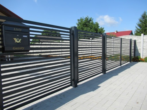 Nowoczesne ogrodzenie posesji z poziomych paneli w kolorze antracytowym, skrzynka pocztowa z napisem 'Listy' i 'Gazety', kostka brukowa na podjeździe.