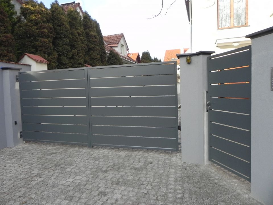 Nowoczesna, szara brama wjazdowa z poziomymi szczelinami, zamontowana na murowanych słupkach, z kostką brukową na podjeździe i zielonymi drzewami w tle.