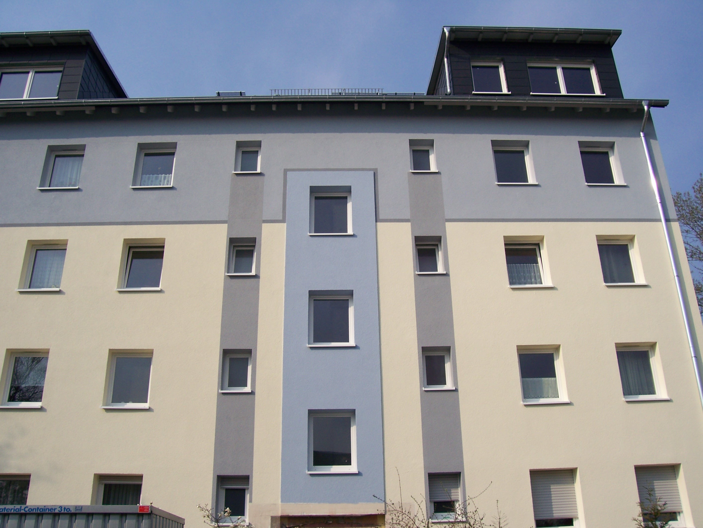 Wielopiętrowy budynek po renowacji z elewacją w odcieniach szarości, beżu i błękitu, z widoczną różnicą kolorów pomiędzy poszczególnymi fragmentami fasady, z oknami z roletami i bez, oraz...