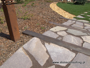 Poligonalne płyty porfirowe ułożone na ścieżce ogrodowej, z widocznymi elementami metalowego obrzeża i żwirową ścieżką w tle, oświetlone naturalnym światłem słonecznym.