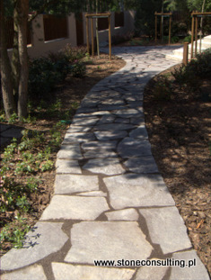 Ścieżka w ogrodzie wyłożona nieregularnymi płytami porfirowymi, prowadząca przez trawnik i nasadzenia w kierunku budynku z drewnianym ogrodzeniem. Widoczne cienie drzew i krzewów.